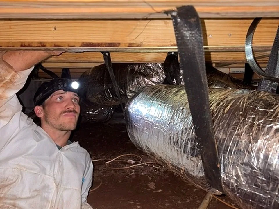 Technician in a white protective coverall and a headlamp working under floor joists in a crawl space, next to insulated flexible ductwork