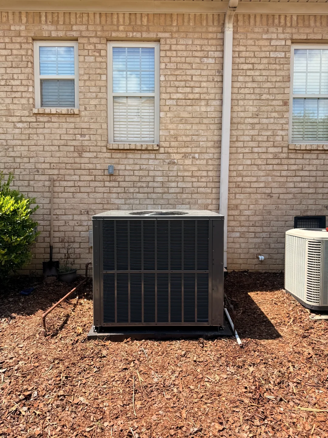 Black packaged HVAC unit against a brick exterior wall next to a smaller outdoor condenser unit