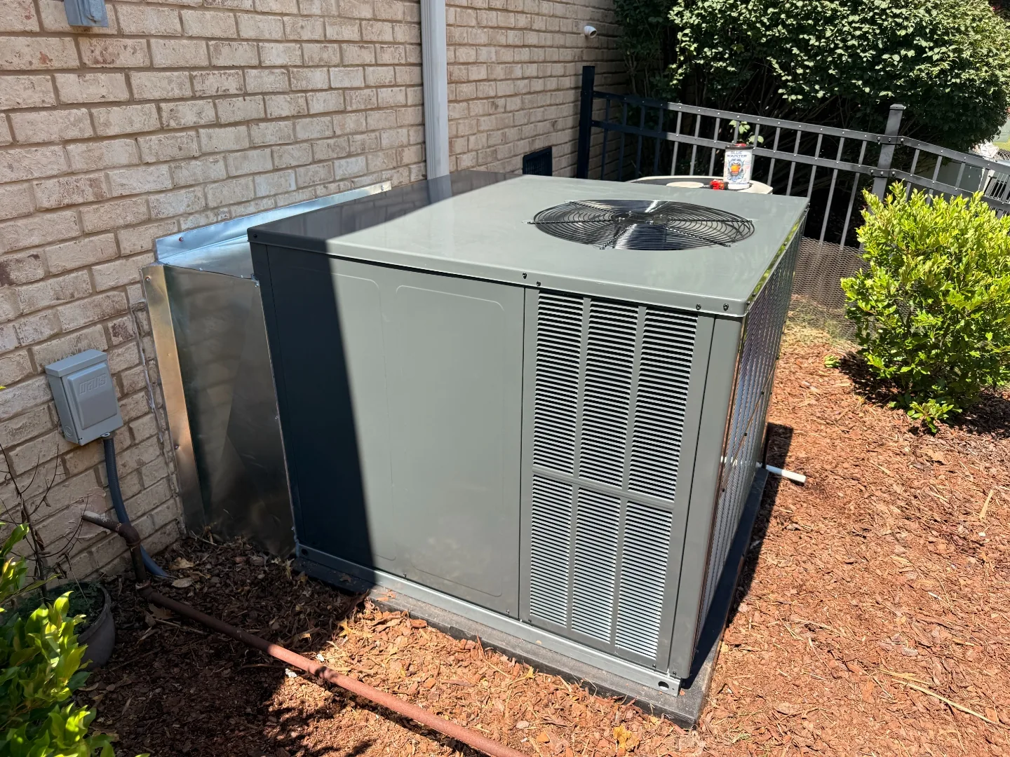 Gray packaged HVAC unit on a concrete pad beside a brick home, with sheet-metal ductwork running up the exterior wall